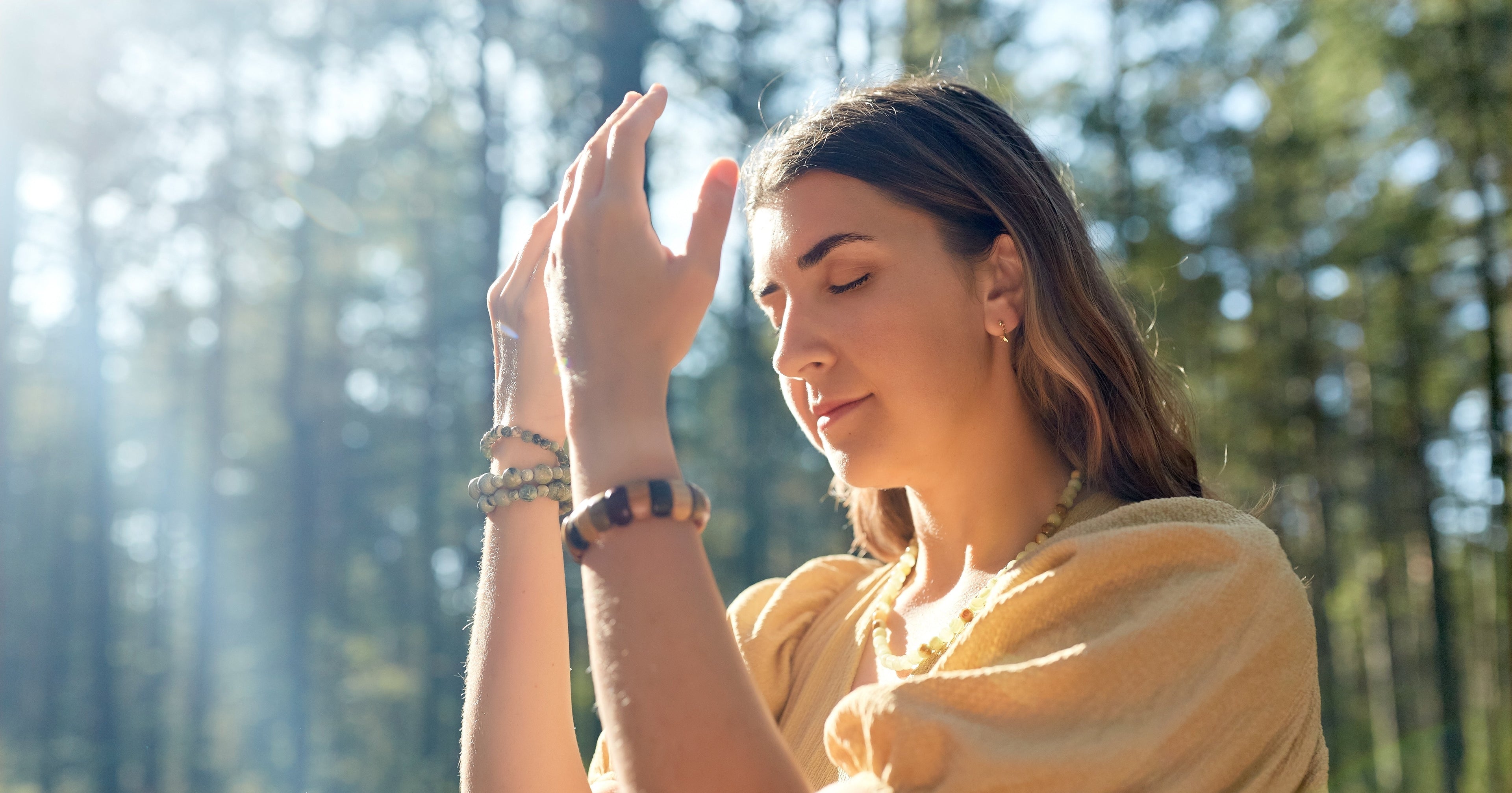 Woman in a yellow dress standing in a forest with her hands raised, eyes closed.